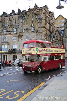 a red double decker bus driving down a street next to tall buildings and people walking on the sidewalk