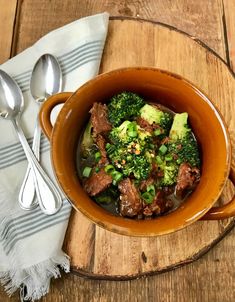 a bowl filled with beef and broccoli on top of a wooden table next to silverware