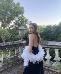 a woman in a black and white feathered dress standing on a stone bridge with trees in the background