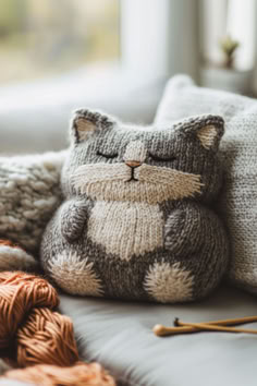 a cat stuffed animal sitting on top of a couch next to yarn and knitting needles