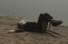 a man laying on the ground next to a chair and body of water in the background