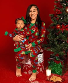 a woman in red pajamas holding a baby next to a christmas tree with presents on it