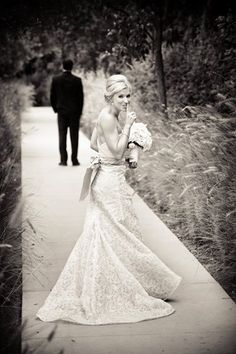 a woman in a wedding dress walking down a path with a man standing behind her