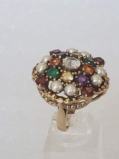 "I'm excited to have come across this beautiful ring because it is in a bigger size and has lots of jewels. The metal is gold plated with a large undercarriage with holes. Each stone is prong set and are in different colored gemstones. The ring has a hallmark \"NB NICKY BUTLER\" and has its original ring box. All stones are intact with non-missing. The ring was used a couple of times and then put away. The finish is shiny with no scratches or other oddities. Makes a great cocktail ring for the H Antique Gold Cluster Ring With Multi-stone, Vintage Multicolor Jewelry With Stone Setting, Vintage Multi-stone Gold Ring, Vintage Gold Multi-stone Cluster Ring, Vintage Gold Multi-stone Ring, Antique Multi-stone Round Jewelry, Antique Cluster Ring As A Gift, Unique Jeweled Rings For Gift, Heirloom Multicolor Ring Jewelry