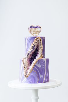 a purple and gold marble cake with a heart shaped top on a white stand against a plain background