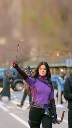 a woman dressed in black and purple holding an arrow while walking down the street with other people