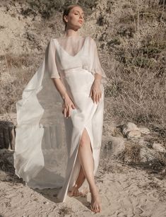 a woman in a white dress is standing on the sand with her hands behind her back