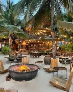 a fire pit surrounded by wooden chairs and tables in front of palm trees on the beach