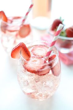 two glasses filled with ice and strawberries on top of a white table next to a bowl full of strawberries