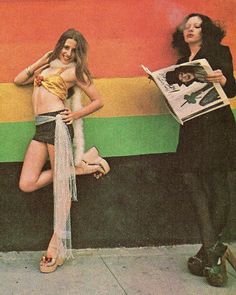 two women standing next to each other in front of a rainbow colored wall with a newspaper