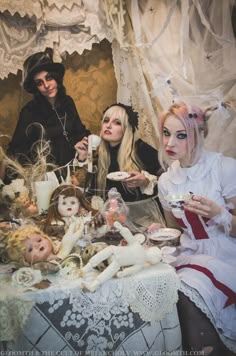 three people dressed up as witches sitting at a table with dolls and food in front of them