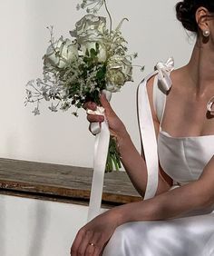 a woman in a white dress holding a bouquet of flowers and sitting on a bench