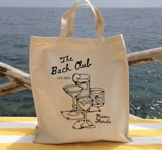 a tote bag sitting on top of a yellow towel next to the ocean with a drink in it