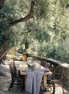 an outdoor table and chairs under trees on the side of a stone wall with water in it