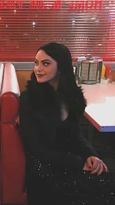 a woman sitting on a red couch in a restaurant with long black hair and makeup