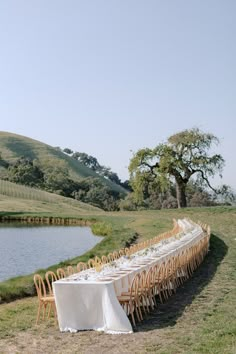 a long table set up with white linens in front of a lake and green hills