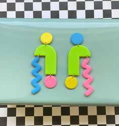 two colorful earrings are hanging on a blue and black checkerboard tablecloth with a green, yellow, pink, and blue design