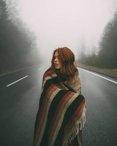 a woman wrapped in a blanket standing on the side of a road with trees behind her