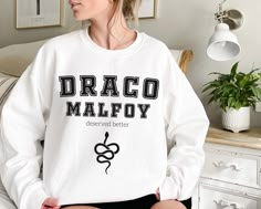 a woman sitting on a bed wearing a white sweatshirt with the word drago malfoy printed on it