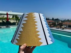 a person holding up a lampshade near a swimming pool with chairs and umbrellas in the background