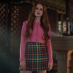 a woman standing in front of a fireplace wearing a pink sweater and multicolored skirt