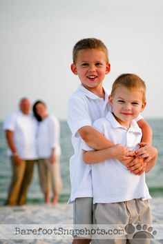 two young boys hugging each other on the beach with their family in the back ground