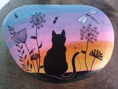 a painted rock with a cat sitting on it's side and flowers in the background