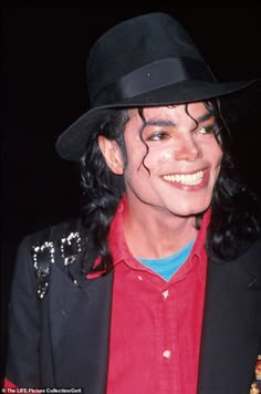 a man with long hair wearing a black hat and red shirt smiles at the camera