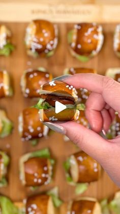 a hand holding a sandwich with lettuce and sauce on it, in front of a tray of sliders