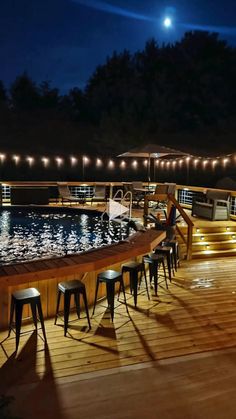 an outdoor deck with bar stools next to a pool at night, lit up by string lights