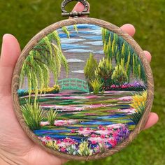 a person holding up a small embroidery art piece in front of a green field with trees and flowers