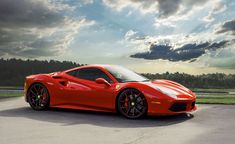 a red sports car is parked on the pavement in front of some trees and clouds