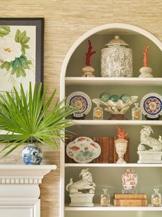 a white book shelf with vases and plates on it next to a potted plant