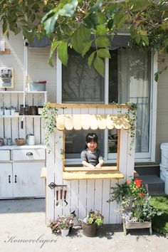 a little boy sitting in the window of a small wooden house with potted plants