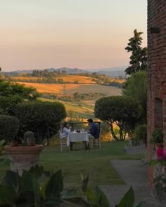 two people sitting at a table in the middle of a garden with rolling hills behind them