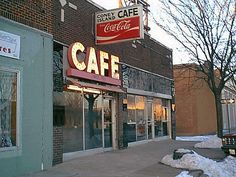 a cafe sign on the side of a building with snow in front of it and cars parked outside