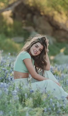 a woman sitting in the middle of a field with blue flowers on her knees and smiling