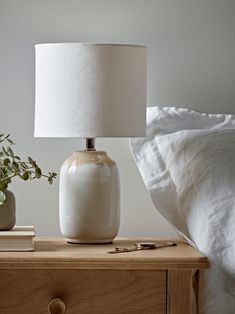 a white lamp sitting on top of a wooden table next to a night stand and pillows