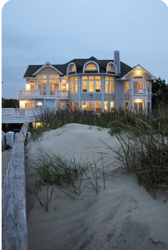 a large house sitting on top of a sandy beach next to a wooden fence with grass growing in front of it