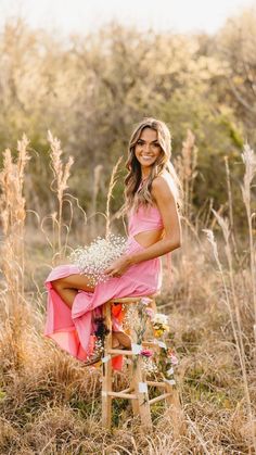 a woman in a pink dress sitting on a wooden stool holding flowers and smiling at the camera