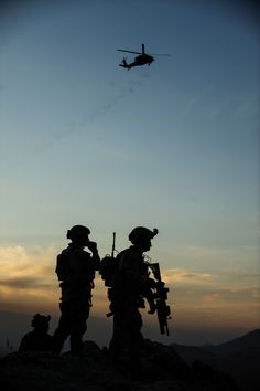 two soldiers standing on top of a hill with a helicopter flying in the sky behind them