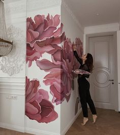 a woman painting a wall with pink flowers on it in a white room next to a chandelier