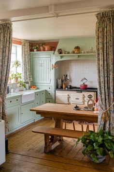 This kitchen combines pastel green cabinetry with floral curtains for a quintessentially English look.

The large wooden table and vintage range are focal points, inviting gatherings and homestyle cooking.

Shelves adorned with potted plants add freshness to the warm atmosphere.