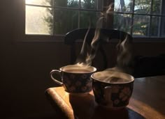 two cups of coffee sit on a table in front of a window, with steam rising from them