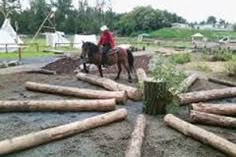 a man riding on the back of a brown horse in a dirt field next to logs