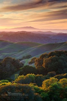 the sun is setting over rolling hills with trees in the foreground and mountains in the background