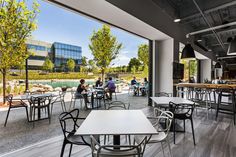 people are sitting at tables in an open area with large windows overlooking the water and trees