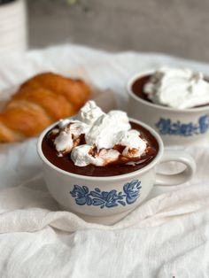 two cups of hot chocolate with whipped cream on top and croissants in the background