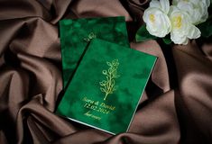 two green wedding guest books on a brown satin background with white flowers in the corner