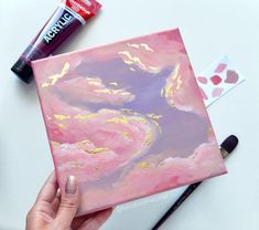 a person holding a pink and gold painting on top of a white table next to paint supplies